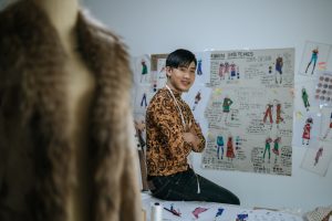 Portrait of Happy Asian young male tailor with measuring tape on neck and looking at camera in fashion studio. Designer and LGBTQ Concept.